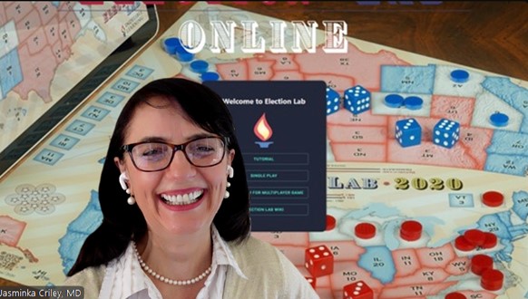 JMC_Oct31stLaugh - Indelible Learning Dr. Jasminka Criley smiling during a video call while presenting Election Lab Online, with a U.S. election-themed board game and dice visible in the background.